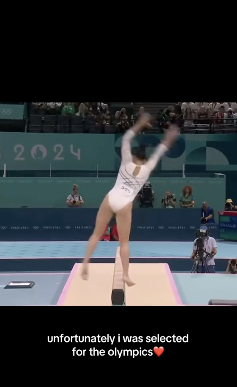 A gymnast in a white uniform on a balance beam performing at the 2024 Olympics, with text below saying, "unfortunately i was selected for the olympics❤️"
