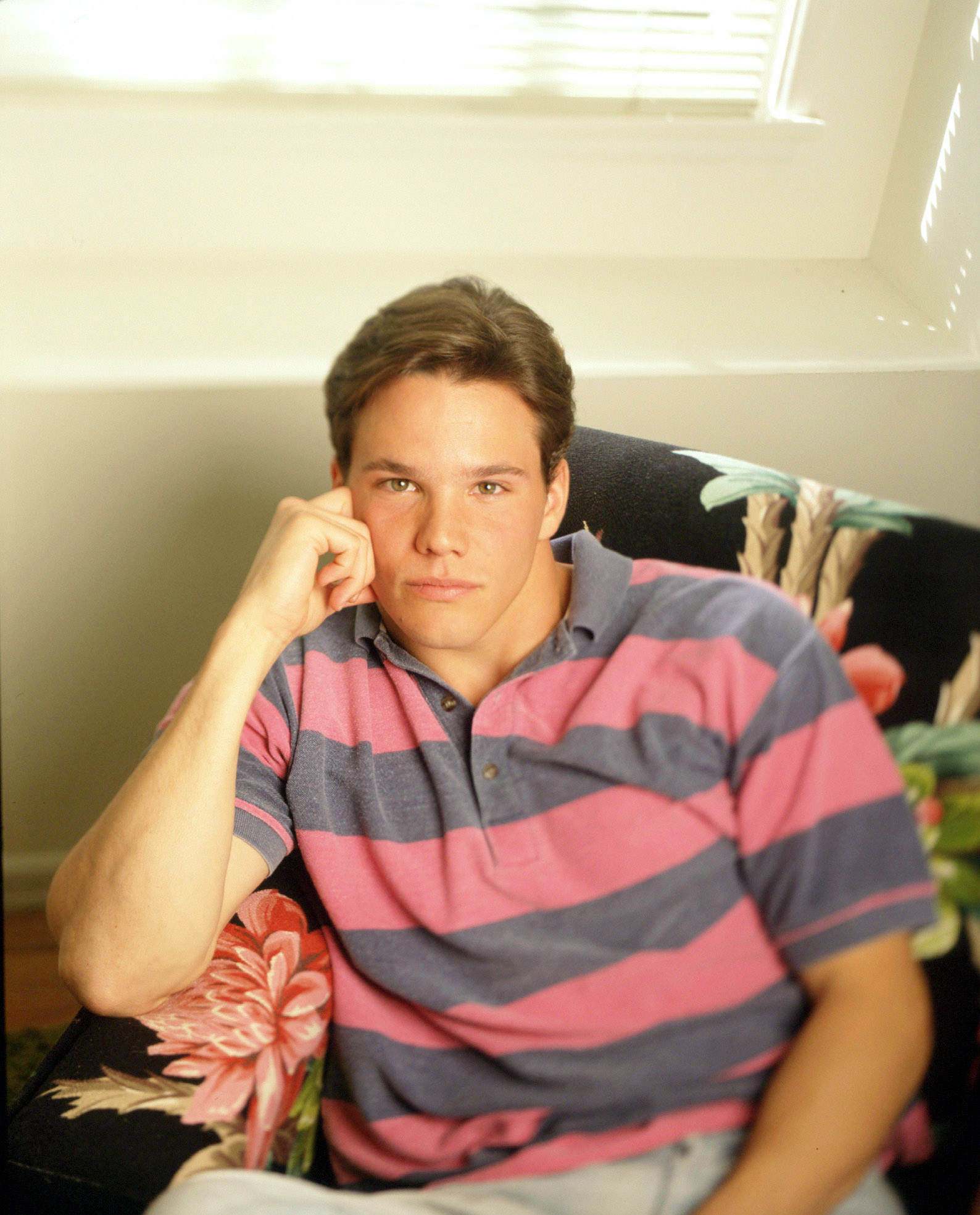 A young Timothy Olyphant lying on a floral-patterned couch wearing a striped polo shirt, resting his head on his hand