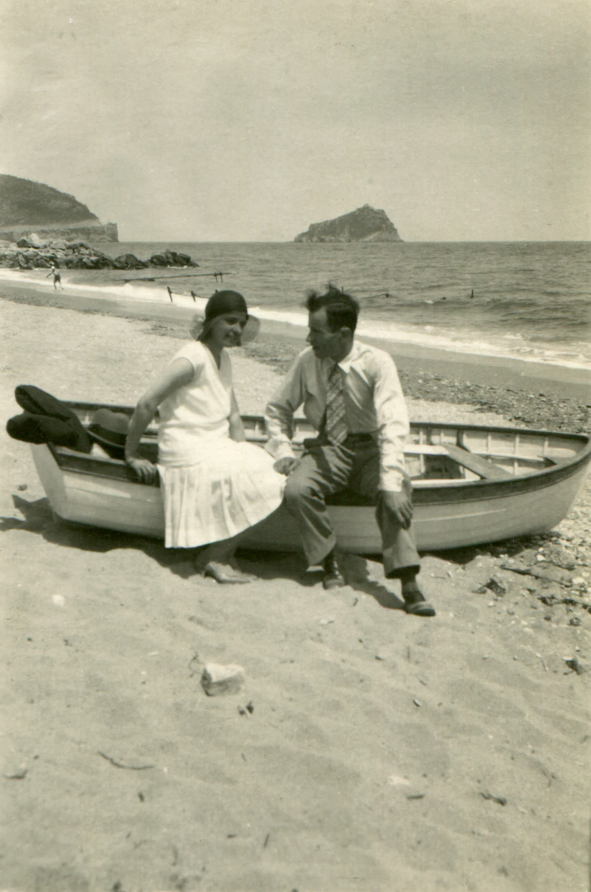 A woman in a sleeveless dress and a man in a shirt and tie sit on a small boat on a beach, with an island and people in the background. Names unknown