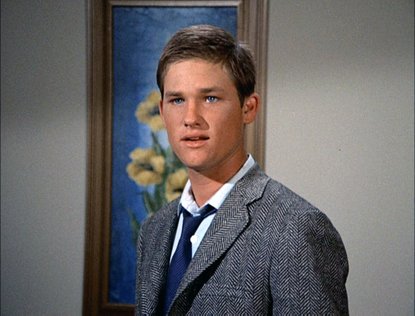 A young man wearing a herringbone jacket, collared shirt, and tie, standing in front of a painting with yellow flowers