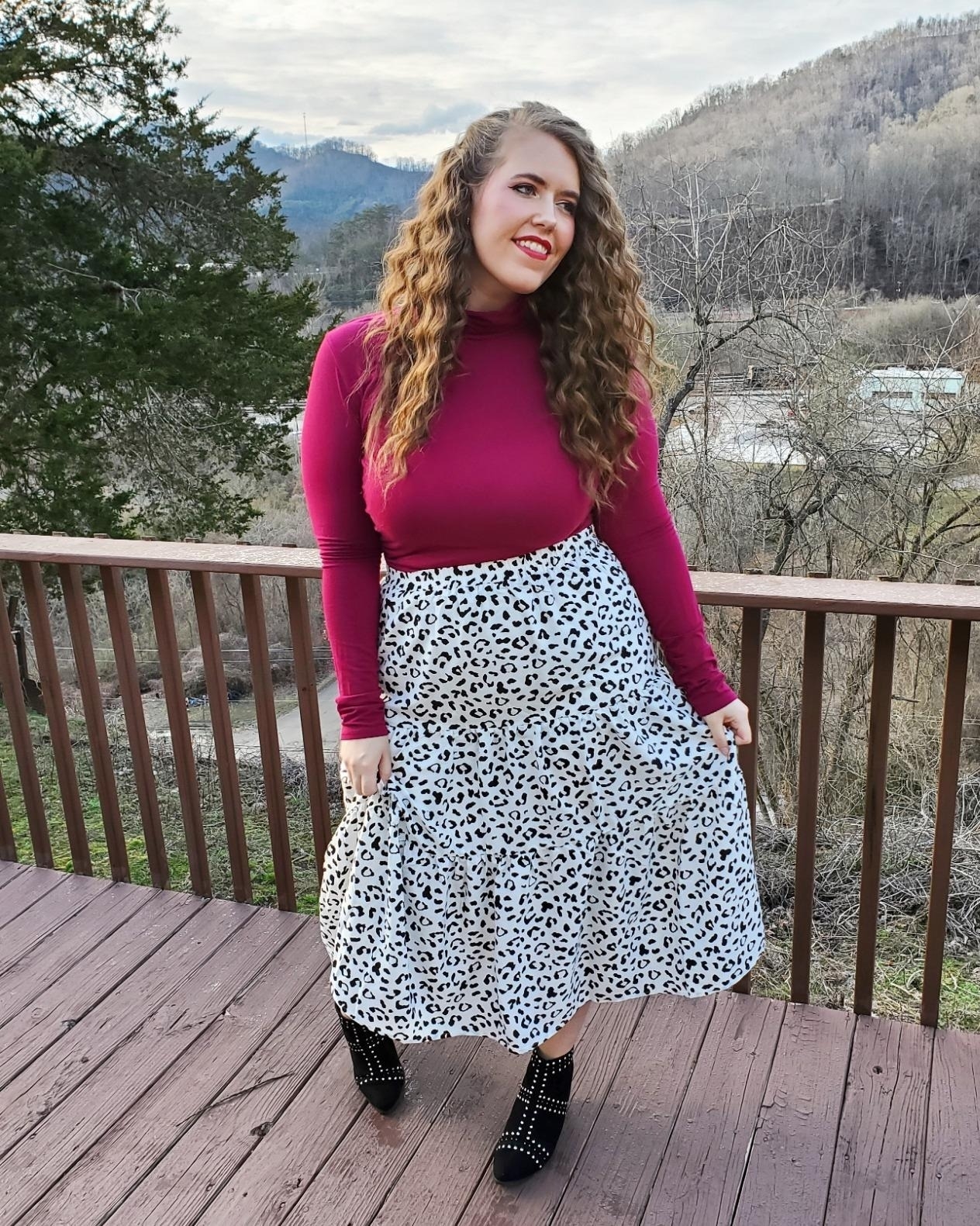 A reviewer wearing a burgundy long-sleeve top and a white animal print skirt stands on a wooden deck with a mountainous backdrop