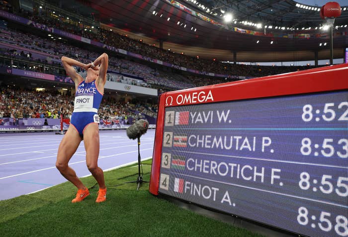 French athlete A. Finot is at the stadium near a scoreboard after a race, appearing emotional. The scoreboard lists race times and competitors: W. Yavi, P. Chemutai, F. Cherotich, and A. Finot