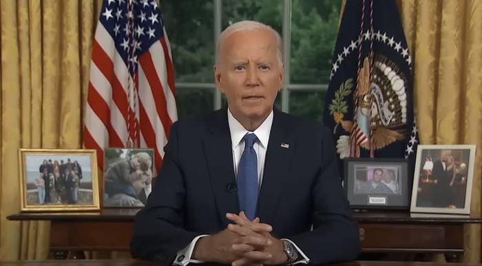 President Joe Biden sitting at a desk with folded hands, delivering a speech. Framed family photos and U.S. flags are in the background