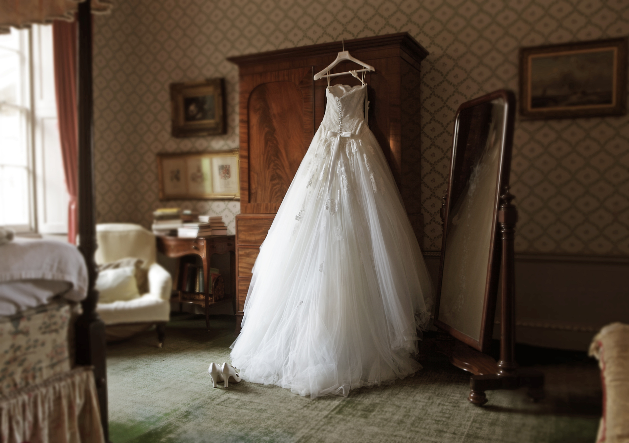 A wedding dress hangs on a wardrobe door in a vintage-style bedroom with a full-length mirror, a pair of shoes on the floor, and assorted furniture