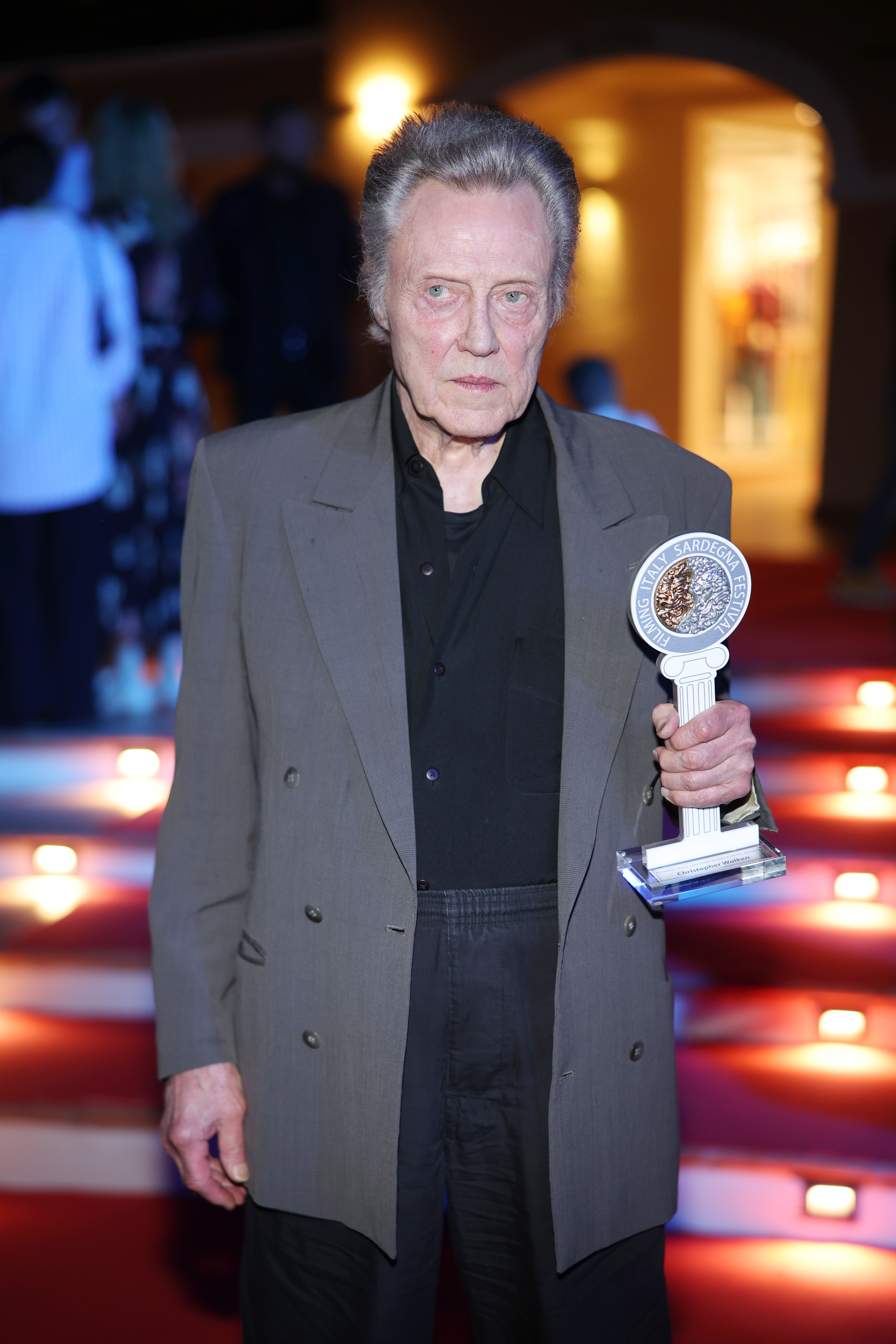 Christopher Walken holding a trophy while standing on a red-carpeted staircase