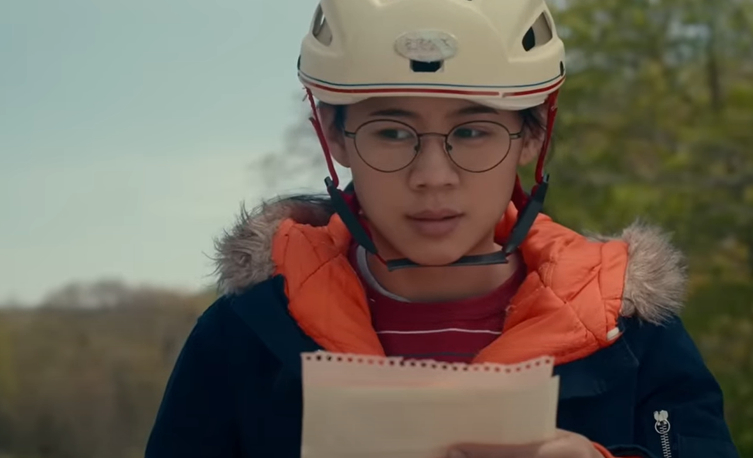 A young girl wearing glasses and a helmet reads a document outdoors
