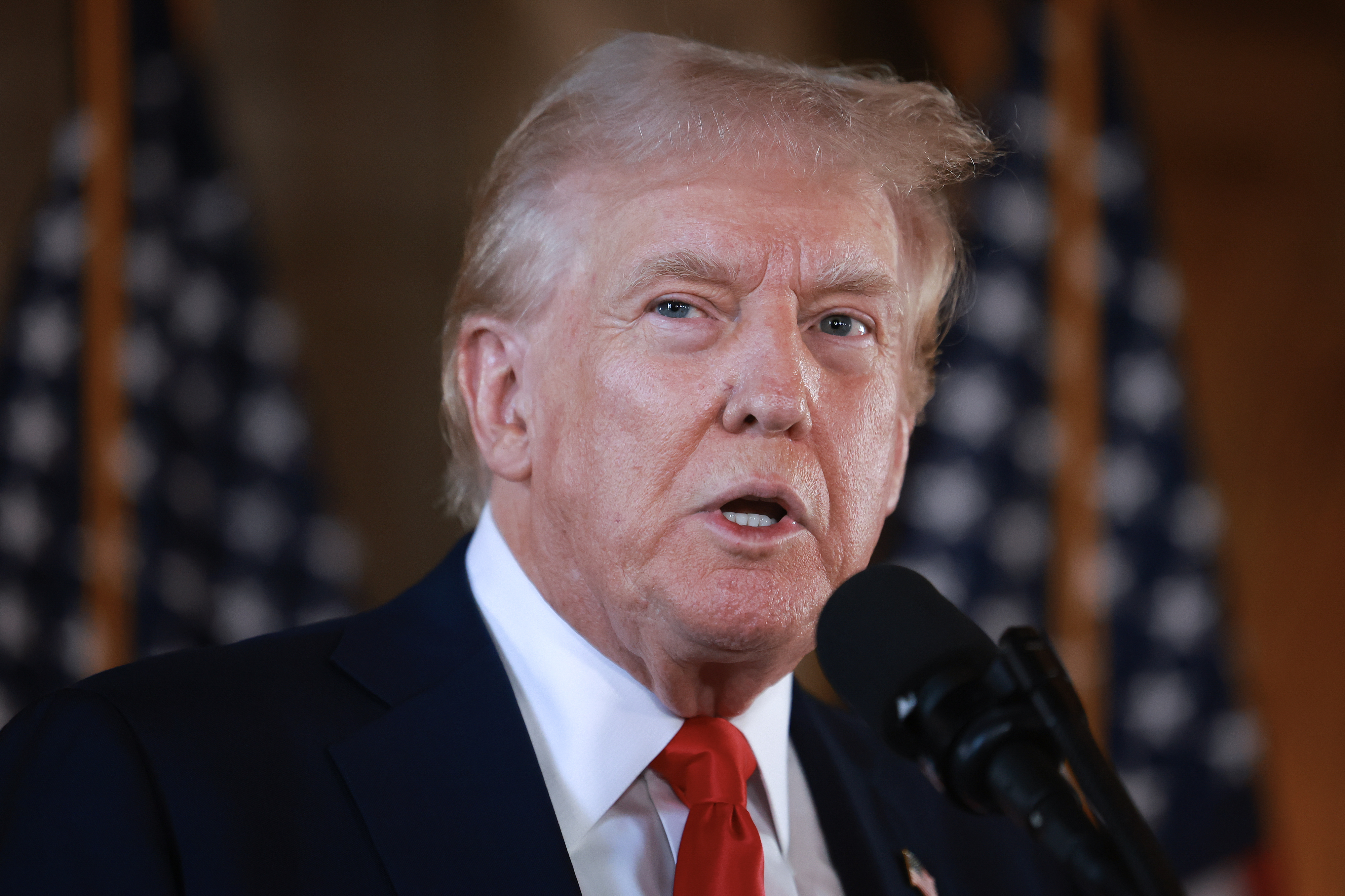 Donald Trump speaking at a podium with American flags in the background. He is wearing a suit and tie