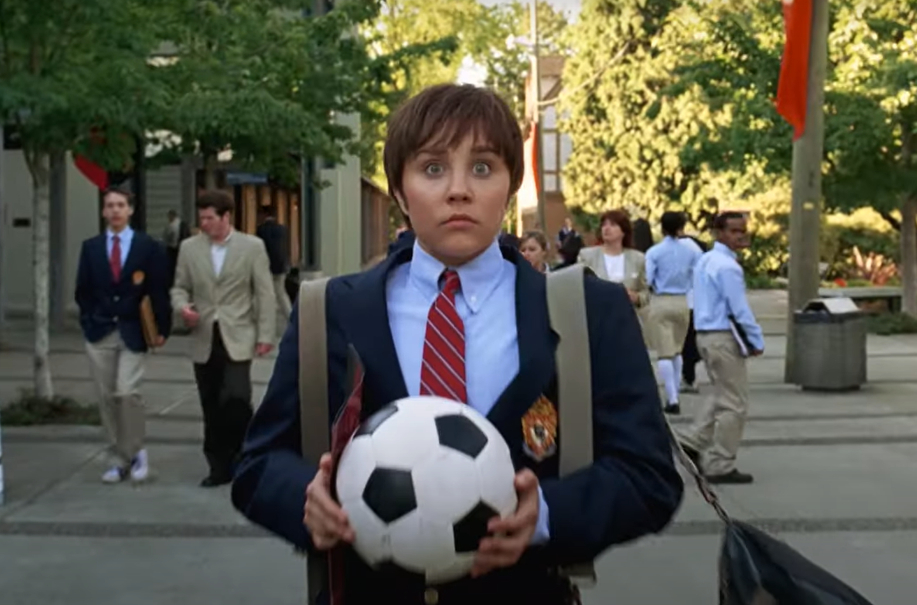 Amanda Bynes in a school uniform holding a soccer ball, looking surprised. She is standing outdoors with other people walking in the background