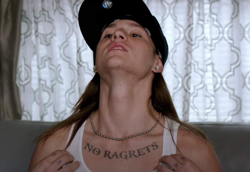Man wearing a black cap and white tank top shows a neck tattoo that reads "NO RAGRETS" with misspelling