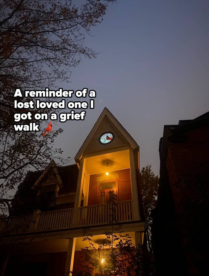 A house with a porch light on at dusk. A circular stained glass window above the porch is illuminated, featuring a colorful pattern. Trees and leaves are partially visible