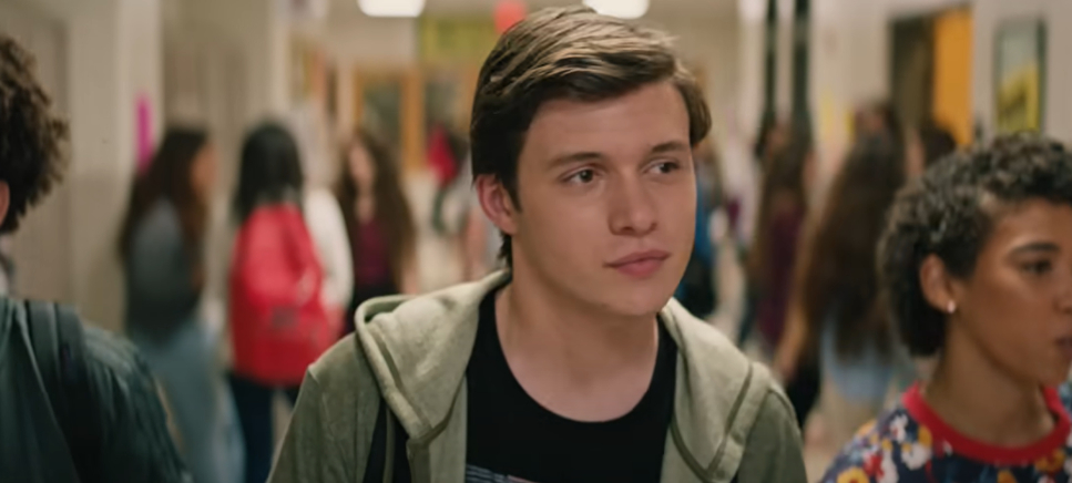 Nick Robinson in a school hallway, wearing a casual jacket and black T-shirt, with other students blurred in the background