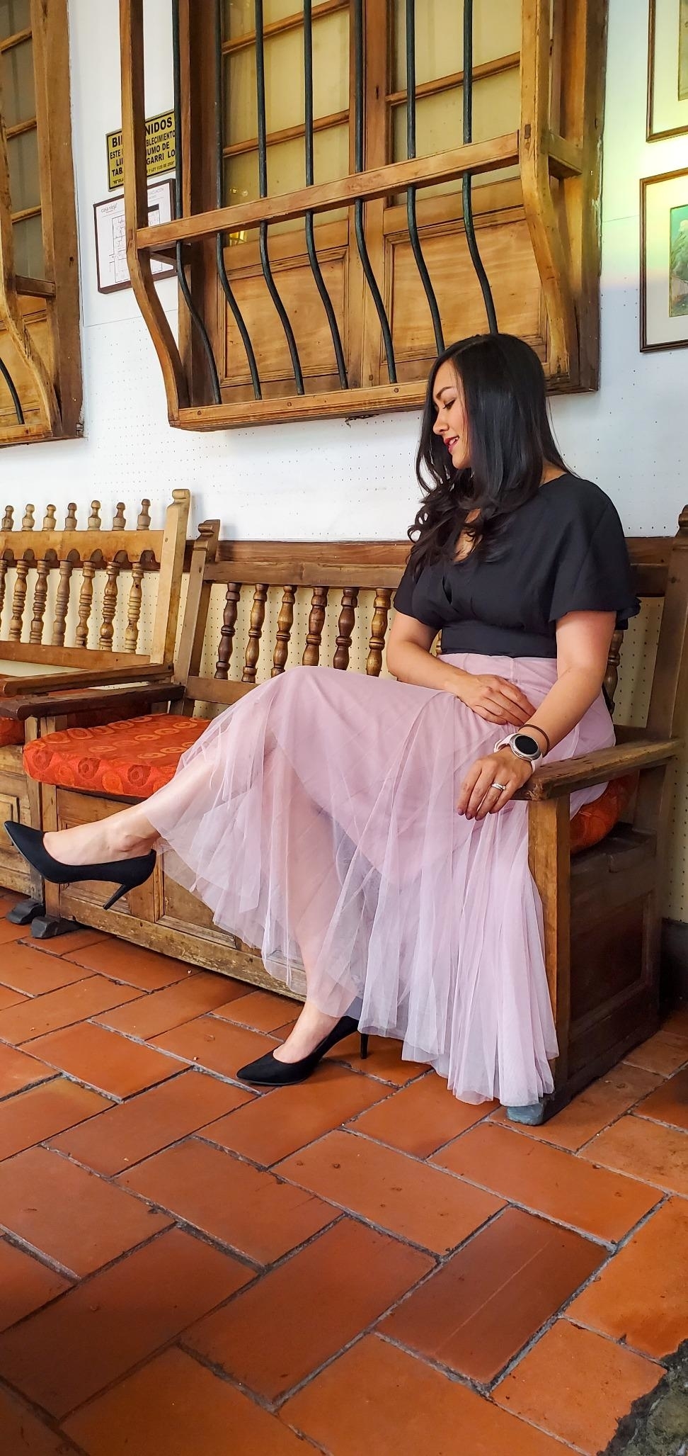 A woman is seated on a wooden bench, wearing a black top and a long, flowing light pink skirt with black heels. The setting appears to be a cozy indoor space