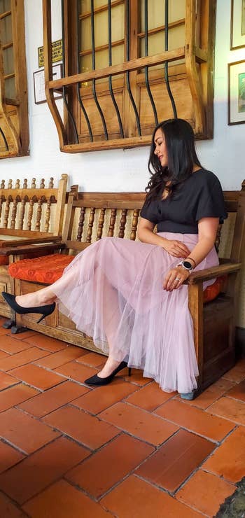 A woman is seated on a wooden bench, wearing a black top and a long, flowing light pink skirt with black heels. The setting appears to be a cozy indoor space