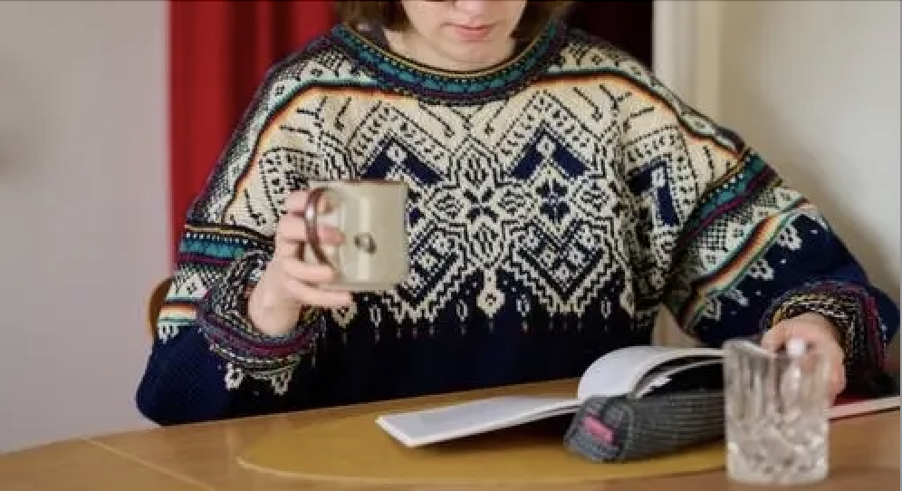 A person in a patterned sweater reads a book at a table while holding a mug. A glass and a pencil case are also on the table