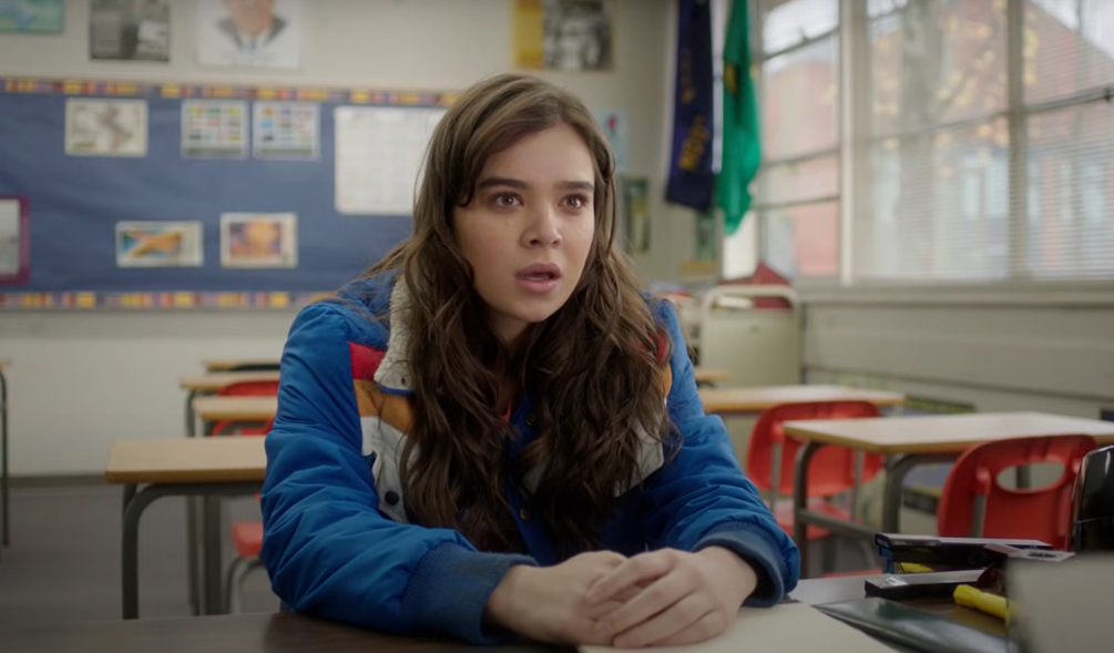 Hailee Steinfeld sits at a classroom desk, looking concerned, wearing a jacket with multicolored stripes in a classroom setting