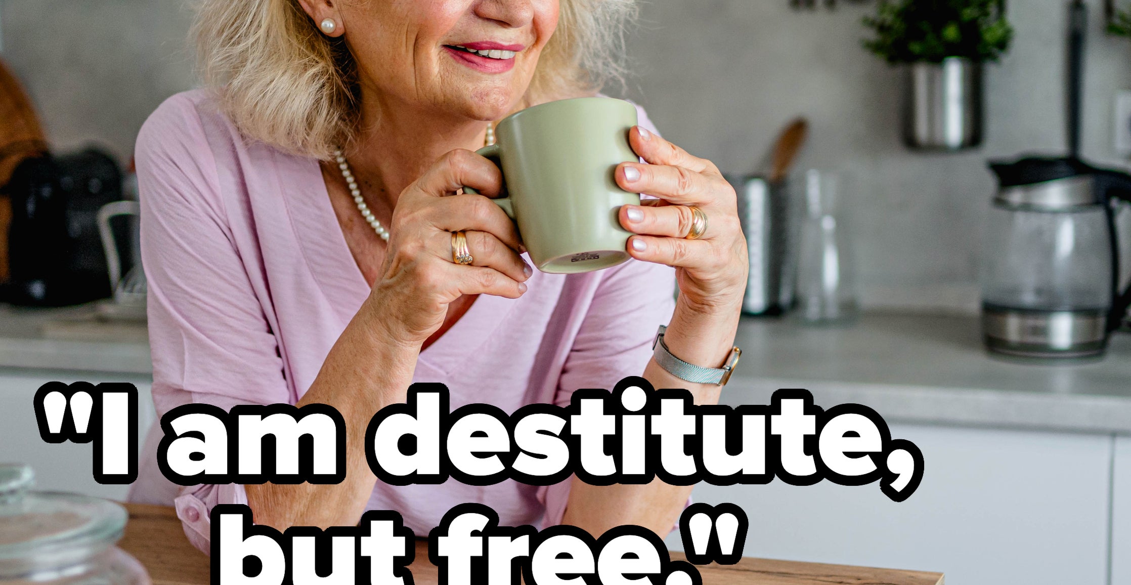 An older woman with gray hair smiles while holding a mug in a modern kitchen, sitting at a wooden counter with a jar of cookies