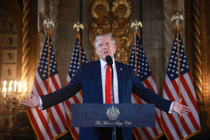 Donald Trump speaks at a podium with "The Mar-a-Lago Club" written on the front, flanked by multiple American flags in an ornate room