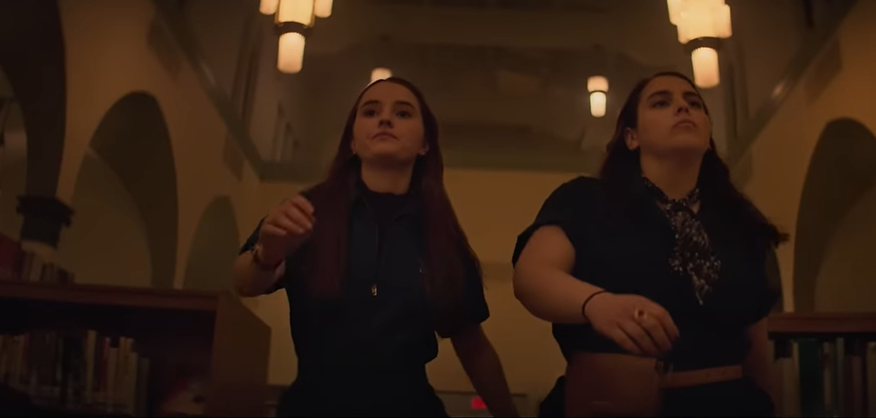 Kaitlyn Dever and Beanie Feldstein are walking through a dimly lit library, looking determined and focused