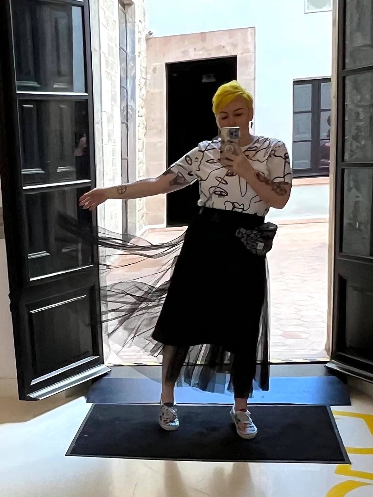 Person with bright yellow hair taking a mirror selfie, wearing a graphic t-shirt, sheer black skirt, patterned socks, and metallic-like shoes, inside a well-lit room