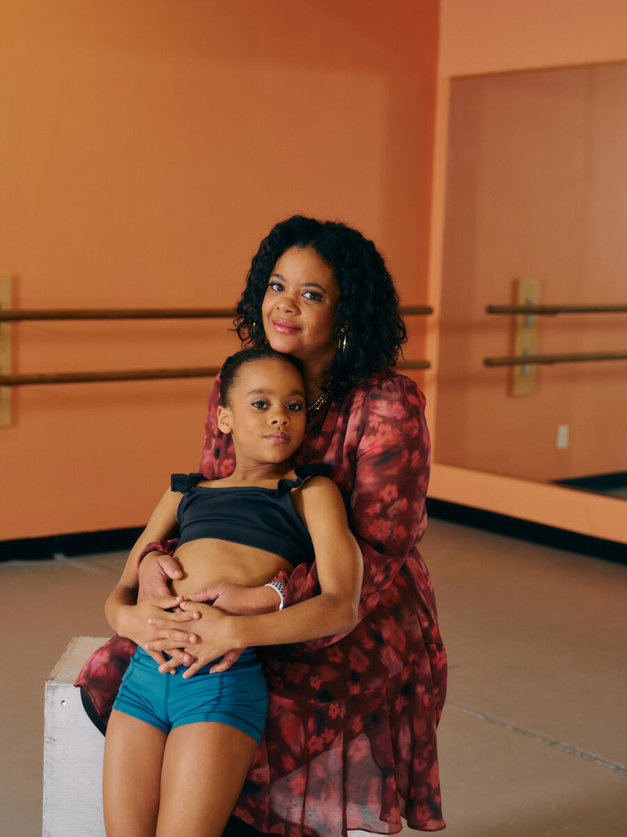 A young girl in dance attire sits on a ballet studio bench, embraced by a woman in a floral dress behind her