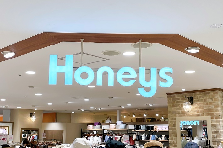 店内の天井から吊り下げられた「Honeys」というブランド名の看板。衣料品が並ぶ売り場が見える。