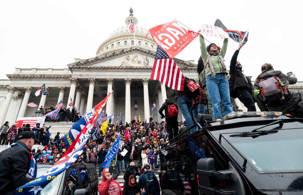Crowds gather in front of the U.S. Capitol building, waving various flags during a political protest. People stand on vehicles and hold signs