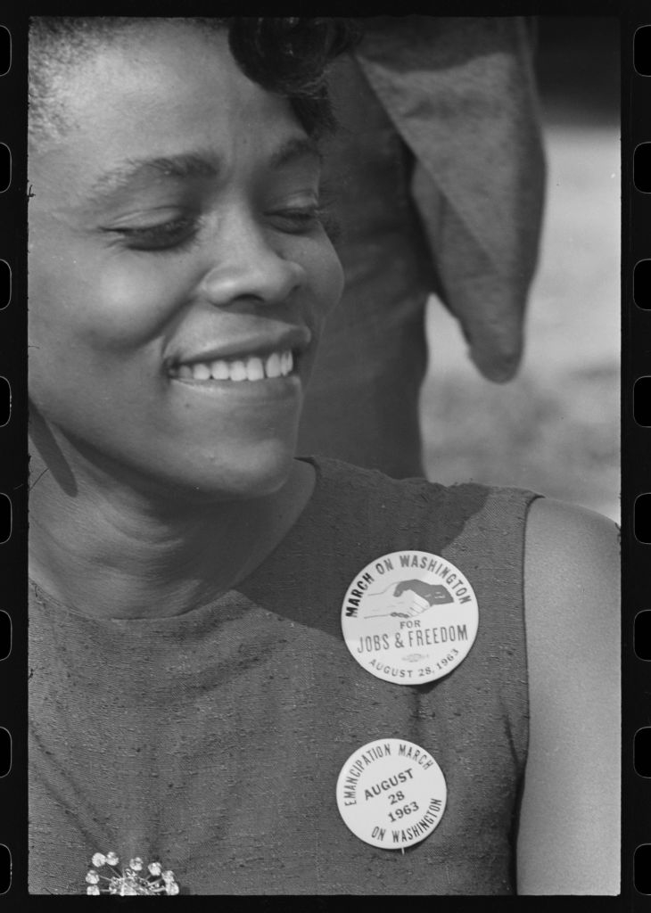 A woman smiles while wearing two buttons reading "March on Washington for Jobs & Freedom August 28, 1963" and "Emancipation March August 1963 on Washington"