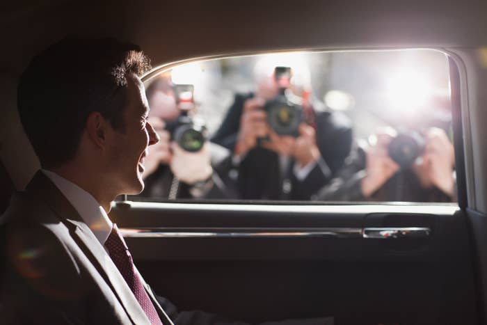 Man in a suit, smiling while sitting in a car, with photographers taking pictures outside the window