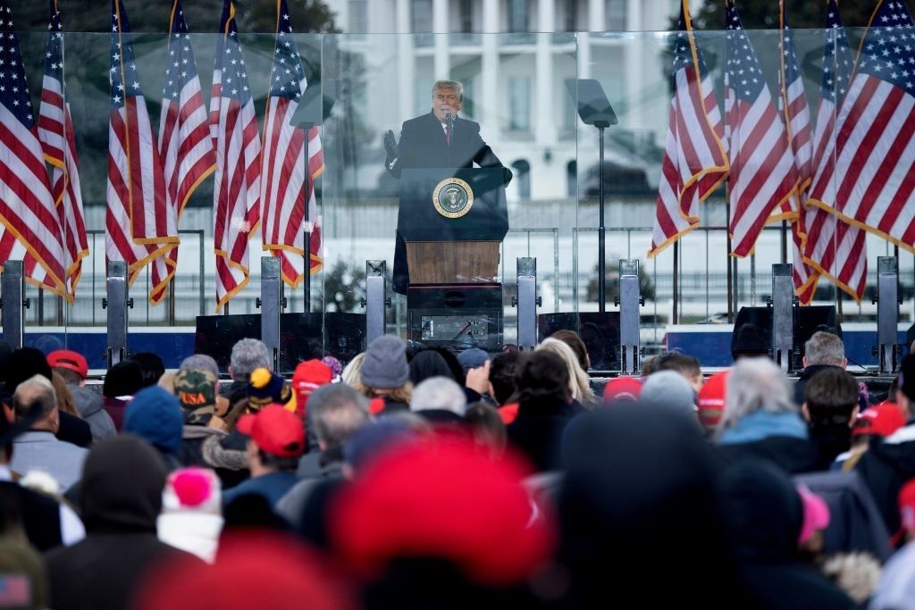 Donald Trump addresses a crowd at a podium with the presidential seal in front of the White House, with numerous American flags displayed around the stage