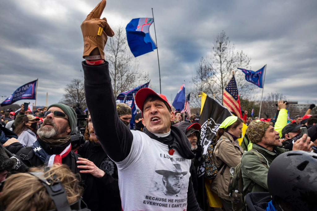 A crowd of people, some waving flags, is shown at a public event. One person in the center is visibly shouting and wearing a shirt with text supportive of Donald Trump