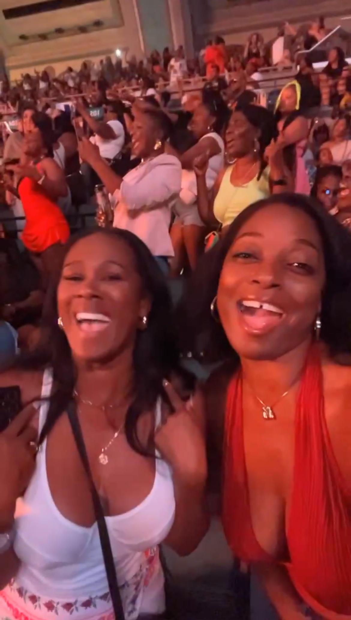 Two unidentified women smile and pose in a lively crowd. They are dressed in sleeveless tops, one in white and the other in red