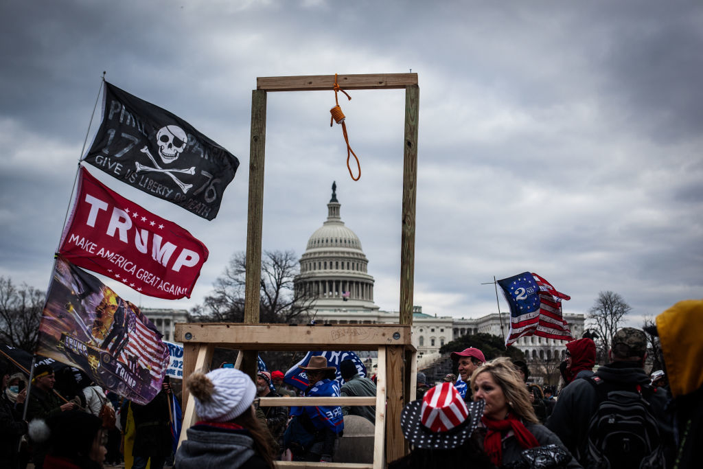 A large wooden gallows with a noose is in front of the U.S. Capitol building, surrounded by people holding flags, including a "Trump Make America Great Again" flag