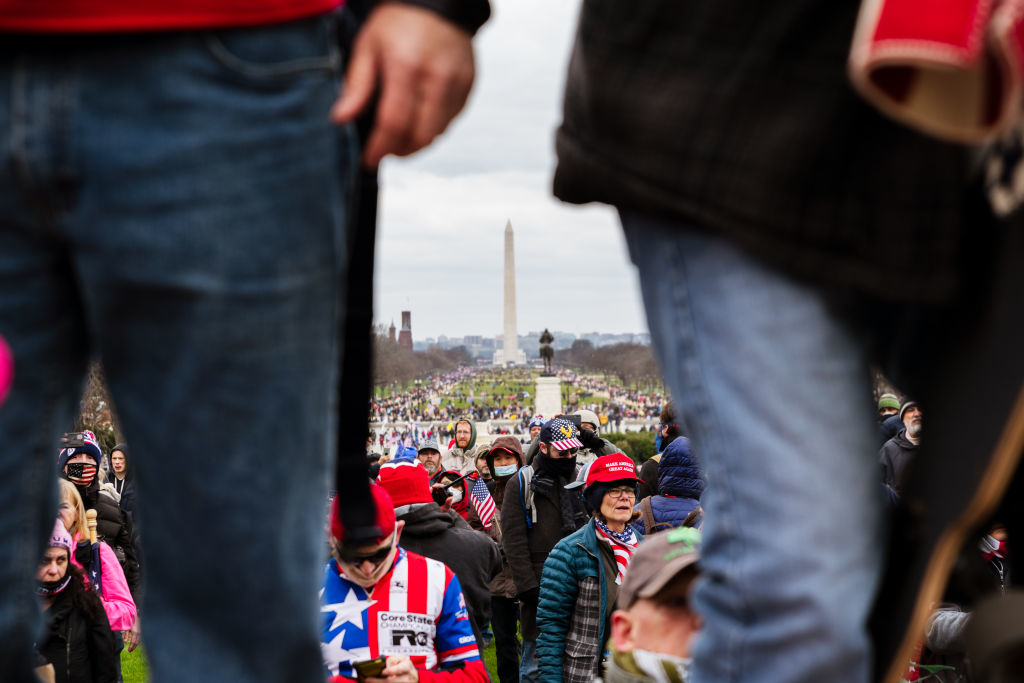 Crowd gathered near the Washington Monument, two individuals in foreground partially obscured, person in red, white, and blue shirt, many wearing hats and flags