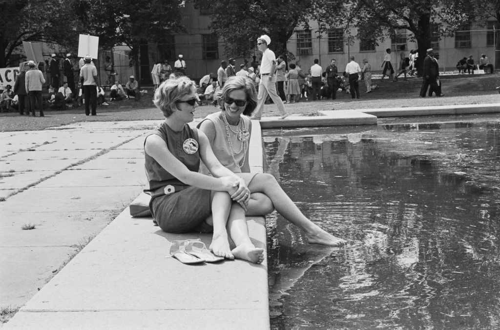 Two women sit on the edge of a pool, chatting and dipping their feet in the water during a public event. People are gathered in the background. Names unknown