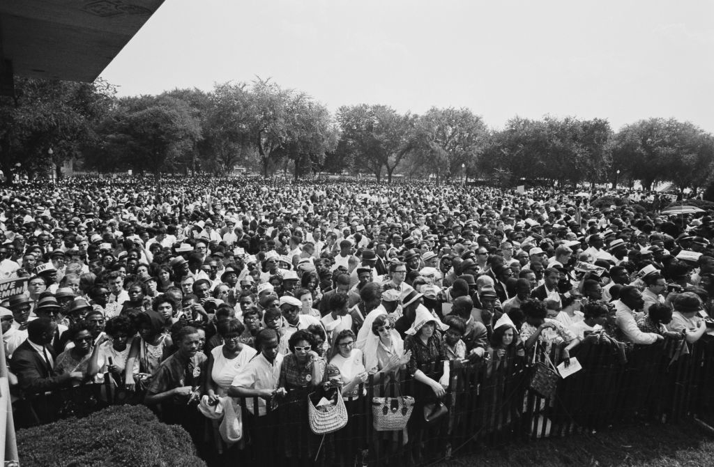 Large crowd of people gathered outdoors for a significant event, many holding signs and wearing summer clothing. Trees are visible in the background