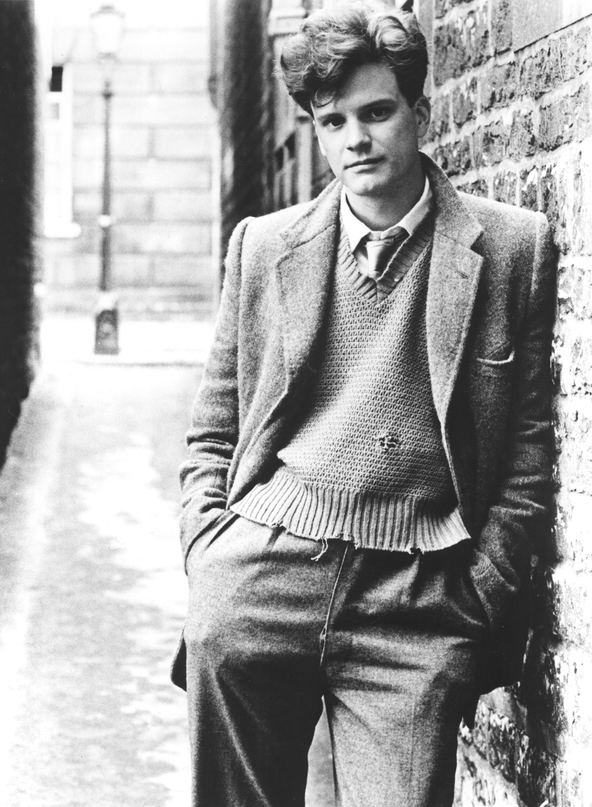 Colin Firth leans casually against a brick wall, dressed in a suit, sweater, and tie