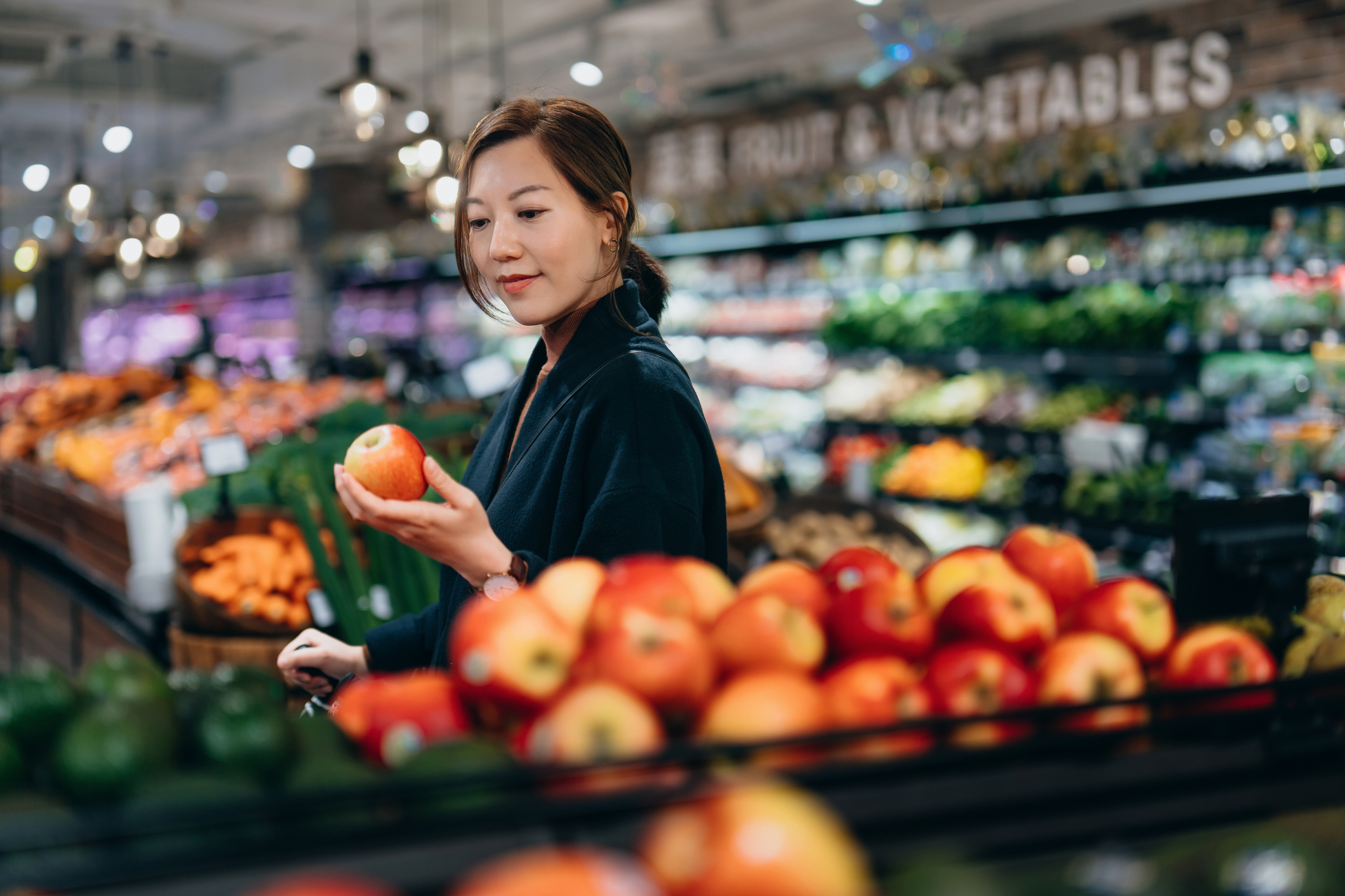 A woman shops for produce in a grocery store, examining an apple. She appears focused on her selection amidst a colorful display of fruits and vegetables