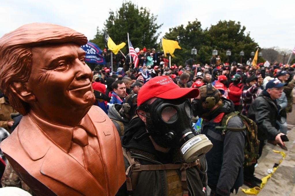 People, some in gas masks and carrying Trump statues, gathered at a protest with flags and trees in the background