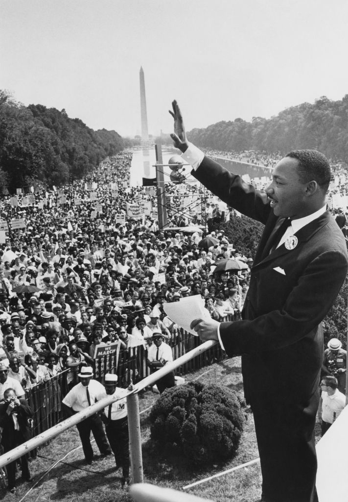 Martin Luther King Jr. delivers a speech, waving to a large crowd gathered at the National Mall, with the Washington Monument in the background