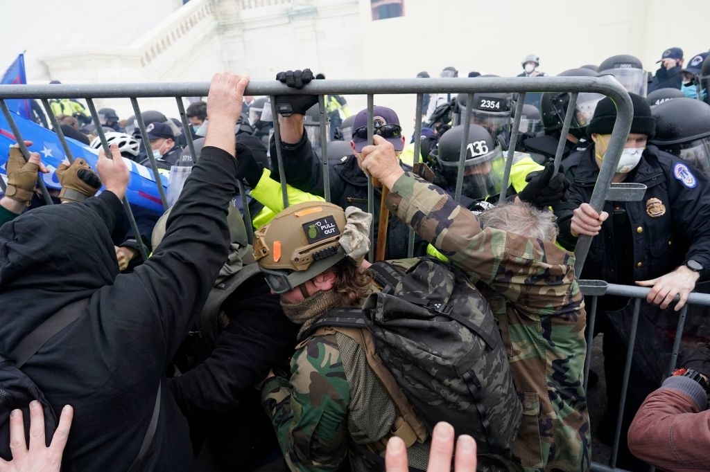 People in tactical gear and police officers clash at a barricade, with some individuals pushing against it. The scene appears chaotic