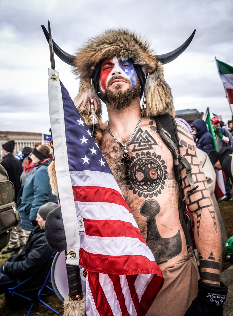 A man, bare-chested with numerous tattoos, wearing a fur headpiece with horns, and face paint, holds an American flag among a crowd outdoors. Names unknown