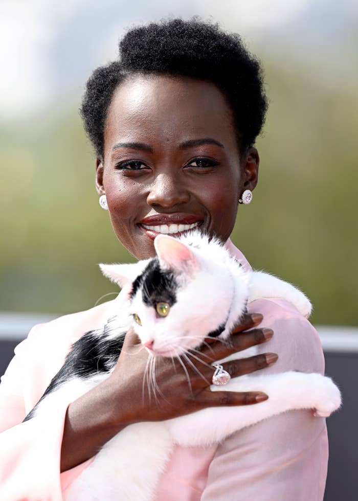 Lupita Nyong'o smiles while holding a black and white cat. She wears a light-colored top and elegant jewelry