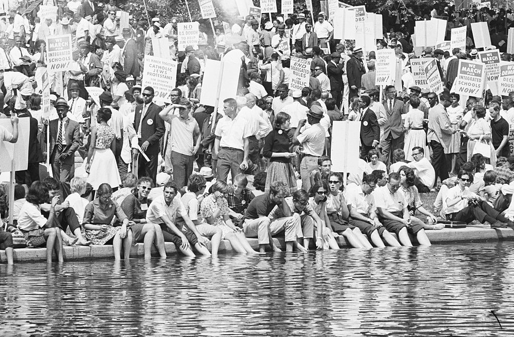 A large, diverse crowd gathered for a civil rights march, with many people carrying protest signs and a group sitting along the edge of a reflecting pool