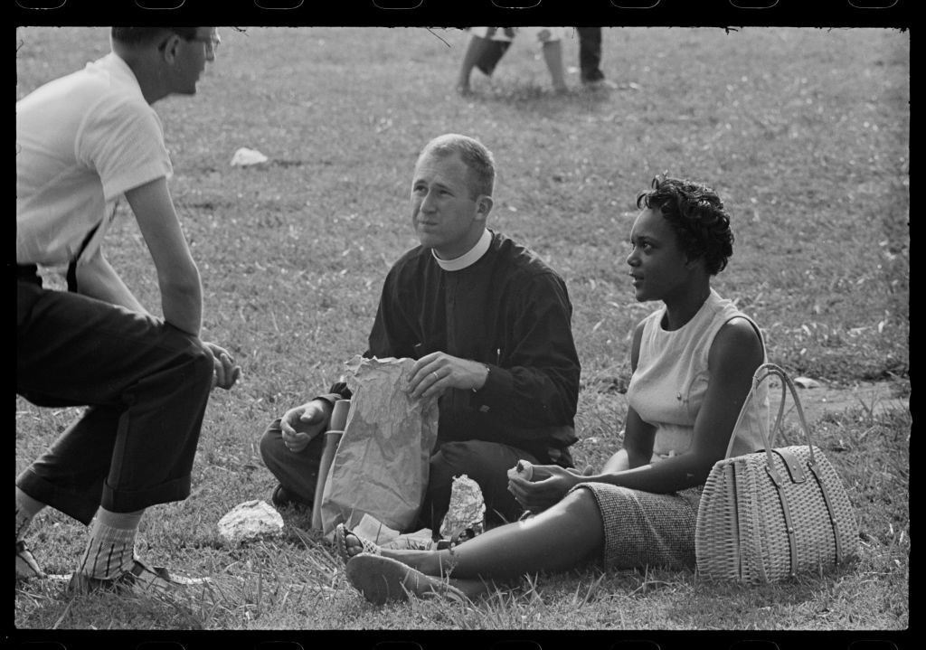 A man, a priest, and a woman sit together on grass, engaging in conversation. The priest holds a paper bag and the woman has a woven bag beside her
