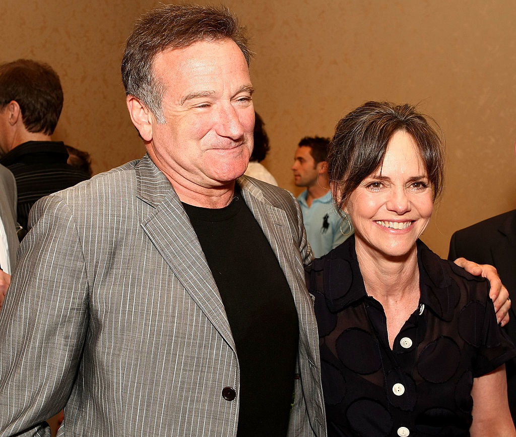 Robin Williams, in a striped suit jacket and black shirt, smiling with Sally Field, wearing a black blouse with a circular pattern, standing beside him