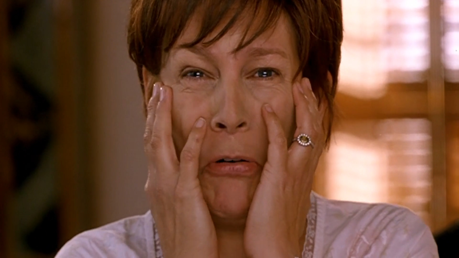 Jamie Lee Curtis looks shocked with hands on her face and mouth slightly open, wearing a light-colored top and a ring on her left hand