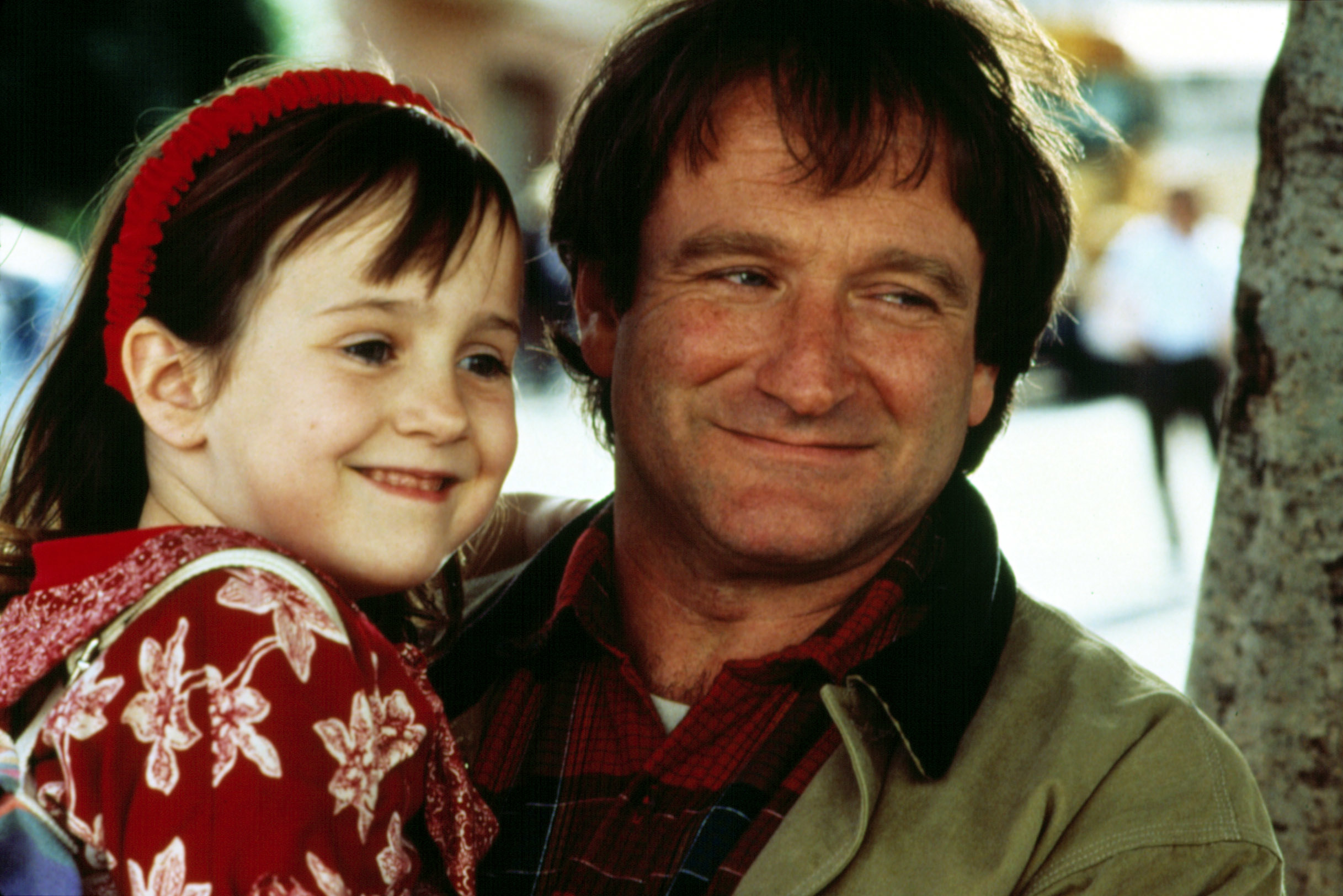 Mara Wilson, in a floral dress and headband, smiles while being hugged by Robin Williams, who is wearing a plaid shirt and jacket