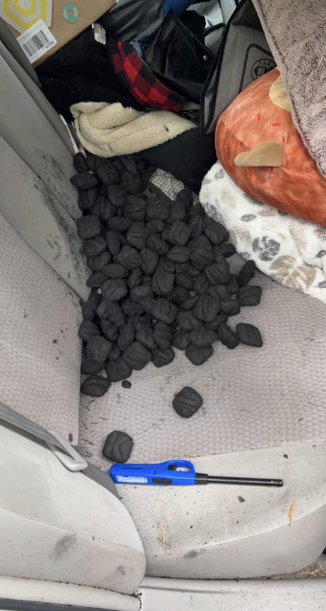 A car's back seat with a pile of black charcoal briquettes scattered on it, a blue lighter, and various blankets and other items surrounding it