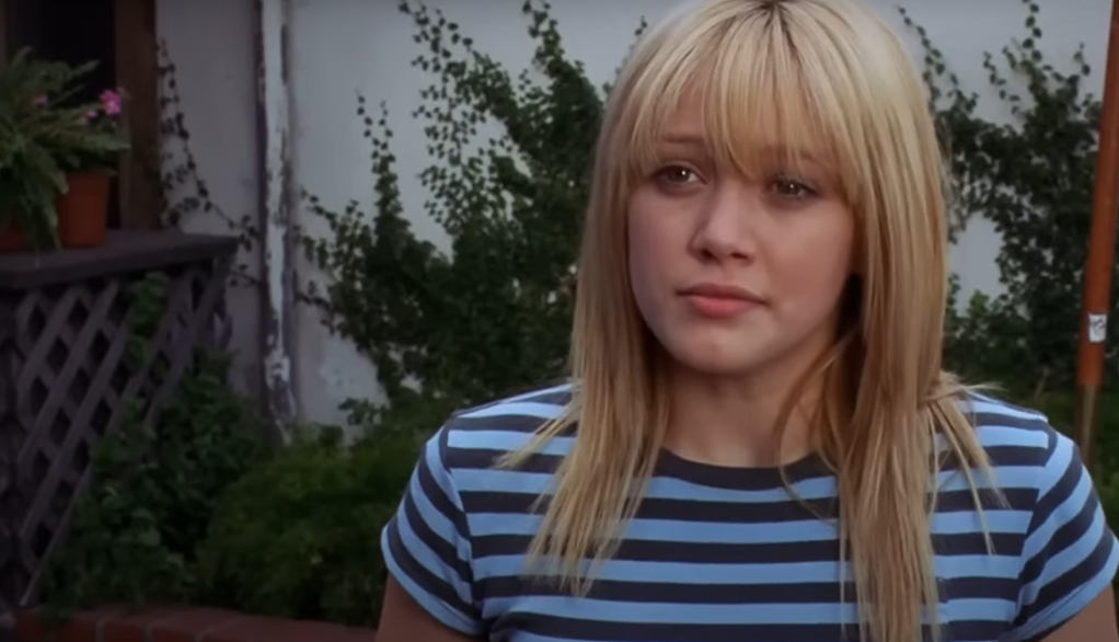 Hilary Duff standing outside with a neutral expression, wearing a short-sleeved, blue and white striped shirt