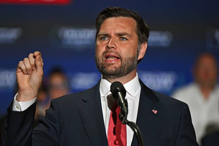 J.D. Vance speaks passionately at a podium during an event, wearing a formal suit and red tie, with a blurred audience in the background